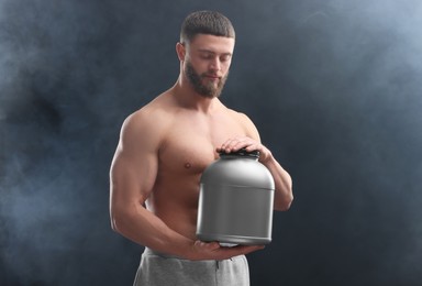 Young man with muscular body holding jar of protein powder on dark grey background
