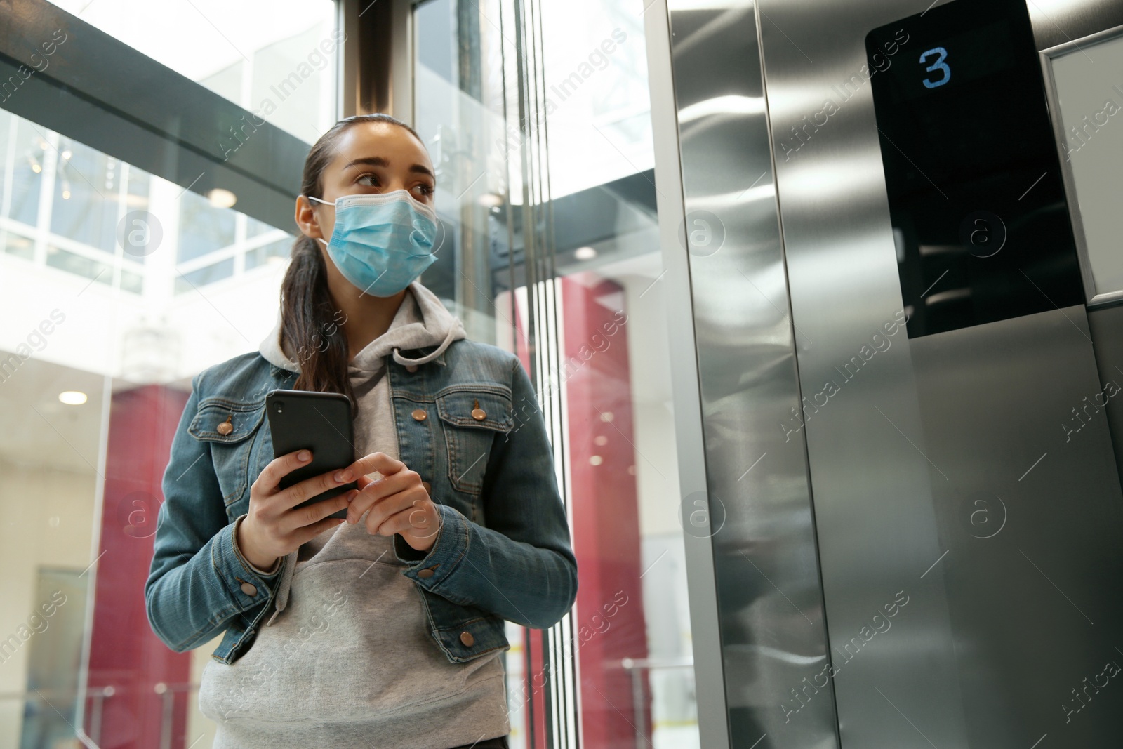 Photo of Young woman with disposable mask and smartphone in elevator. Dangerous virus