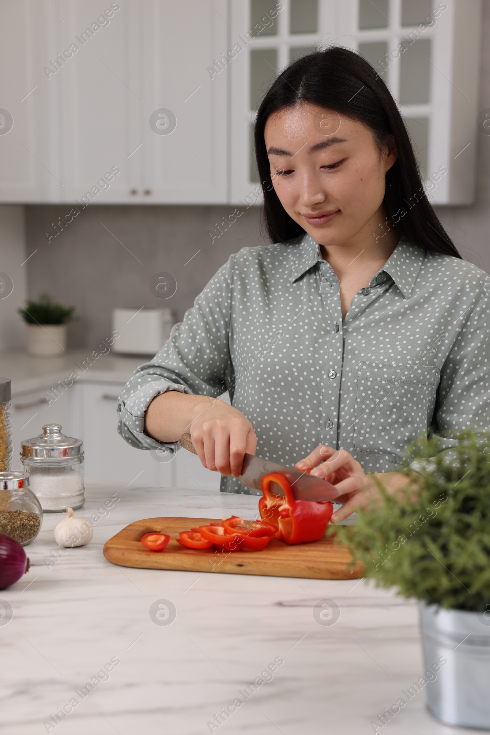 Photo of Cooking process. Beautiful woman cutting bell pepper at white marble countertop in kitchen