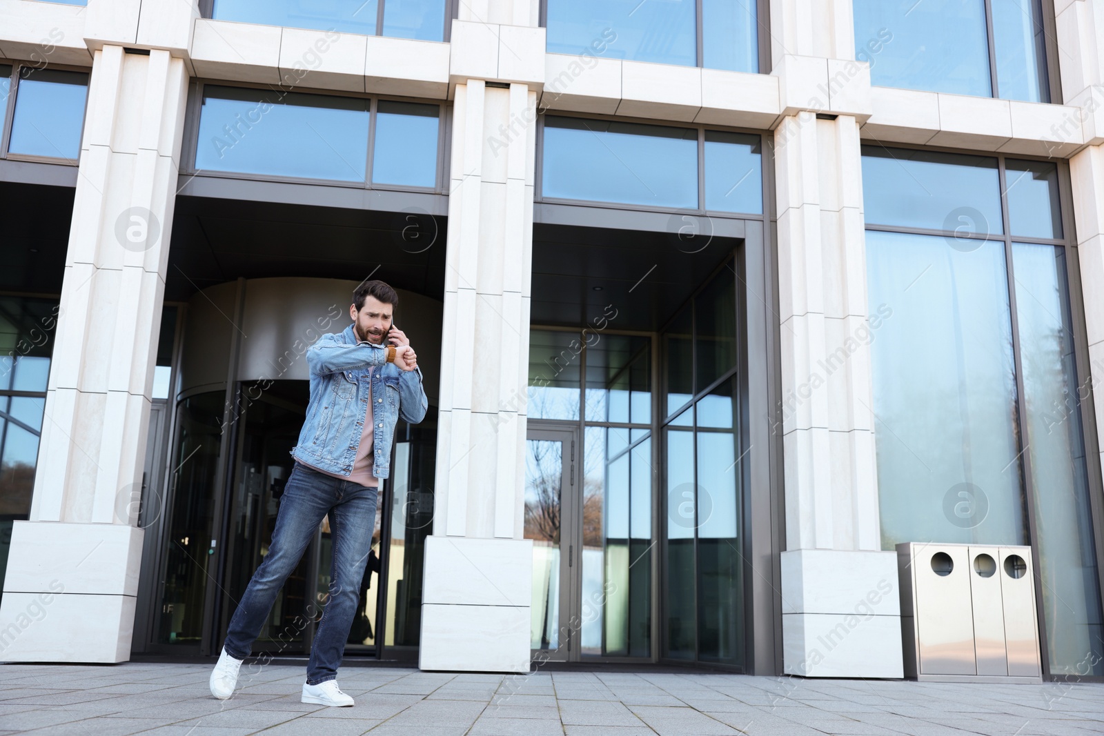 Photo of Emotional man talking on smartphone and looking at watch near building. Being late concept