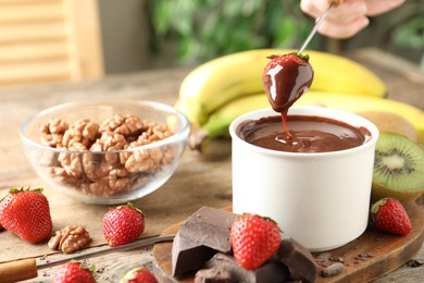 Photo of Woman dipping strawberry into fondue pot with chocolate at wooden table, closeup