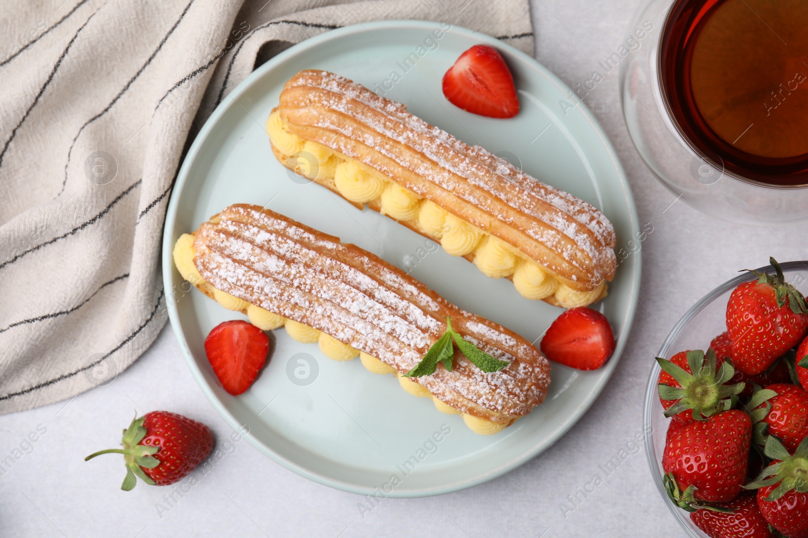 Photo of Delicious eclairs filled with cream, strawberries, mint and tea on light table, top view