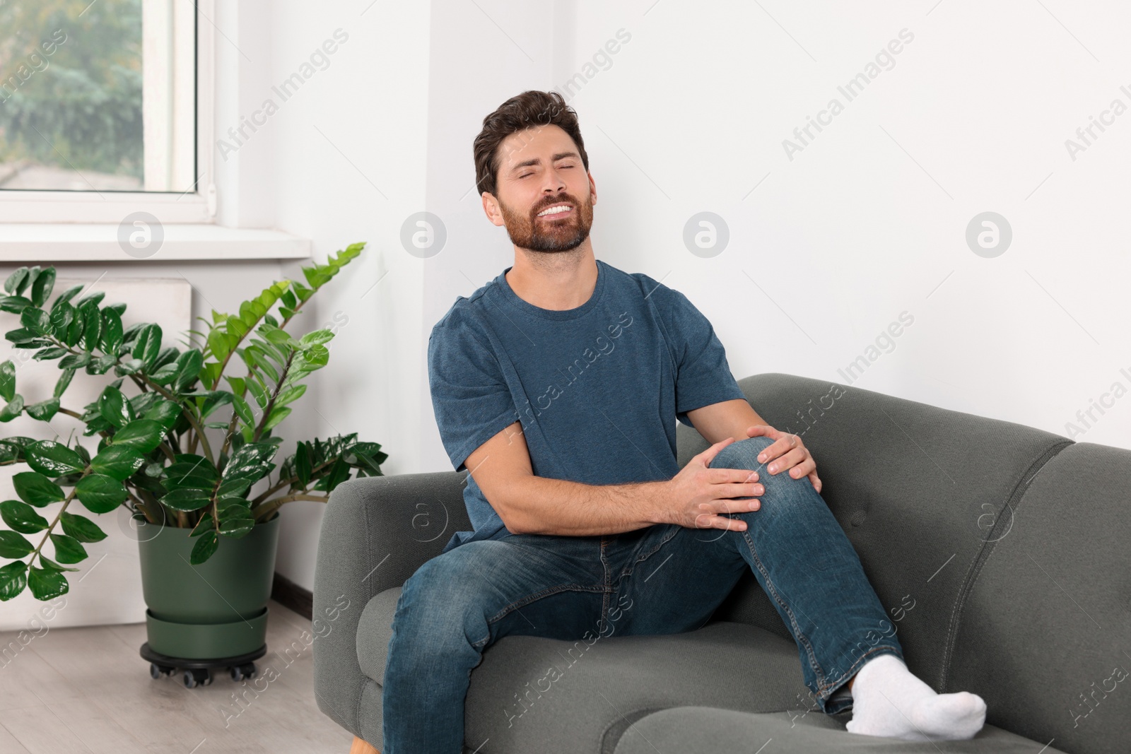 Photo of Man suffering from leg pain and touching knee on sofa at home