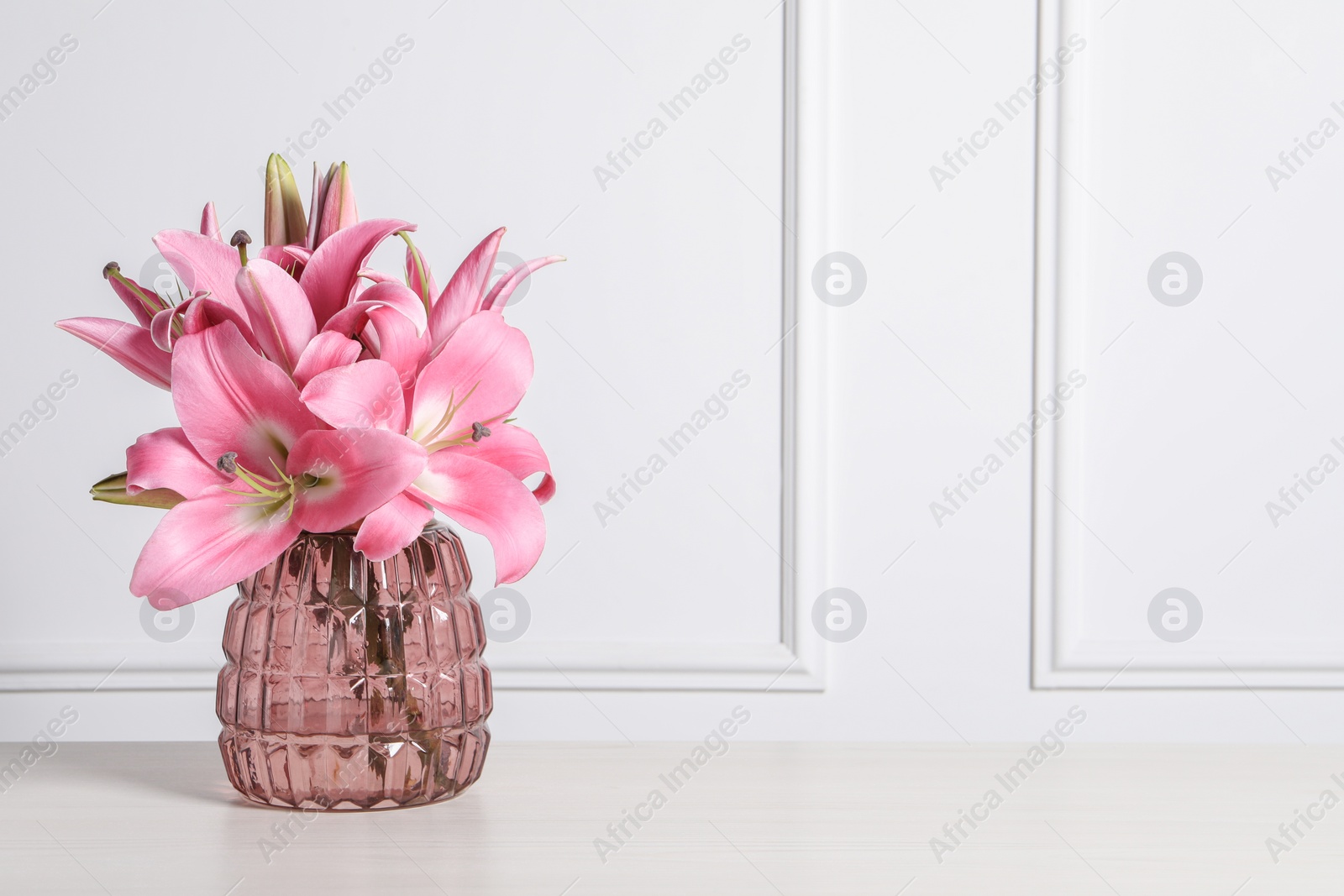 Photo of Beautiful pink lily flowers in vase on wooden table against white wall, space for text