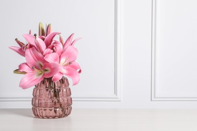 Photo of Beautiful pink lily flowers in vase on wooden table against white wall, space for text