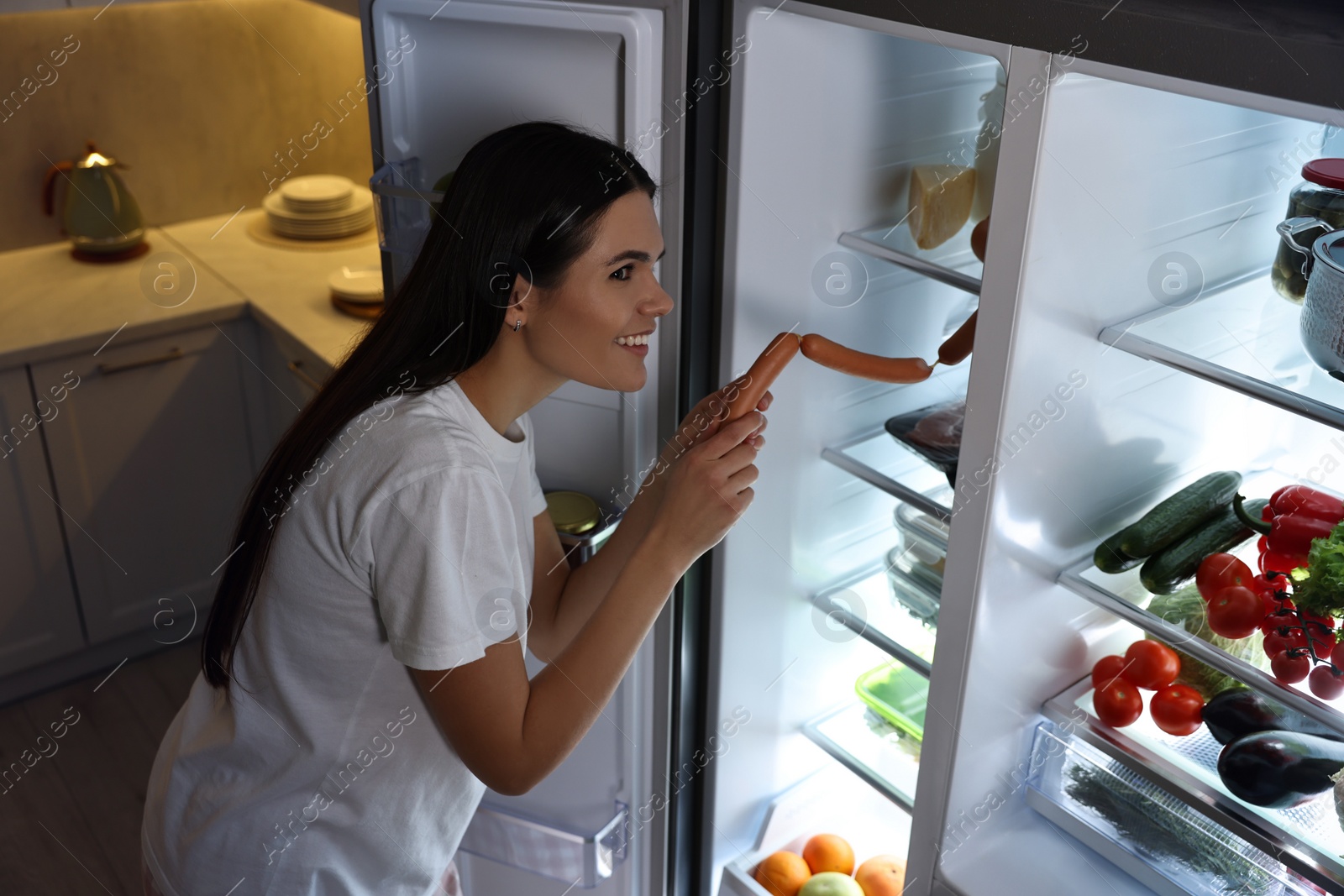 Photo of Young woman taking sausages out of refrigerator in kitchen at night