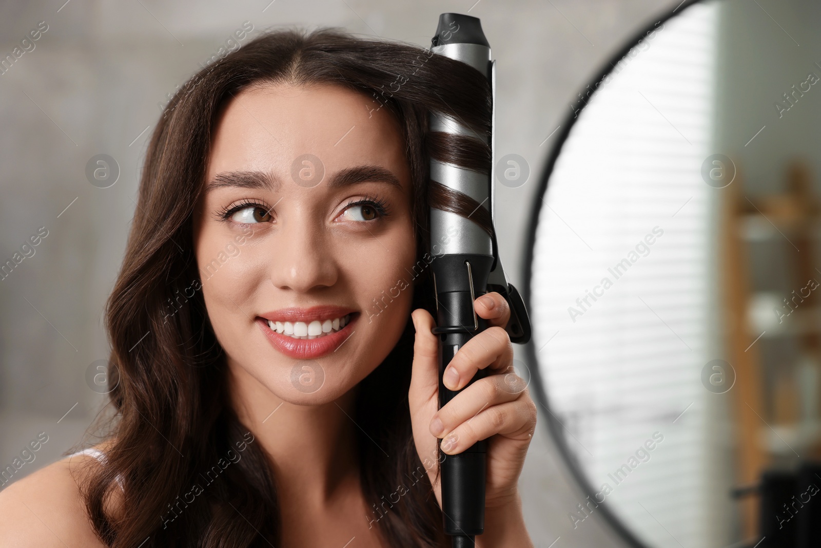 Photo of Smiling woman using curling hair iron in bathroom