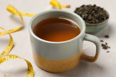 Cup of herbal diet tea and measuring tape on light marble table, closeup. Weight loss concept