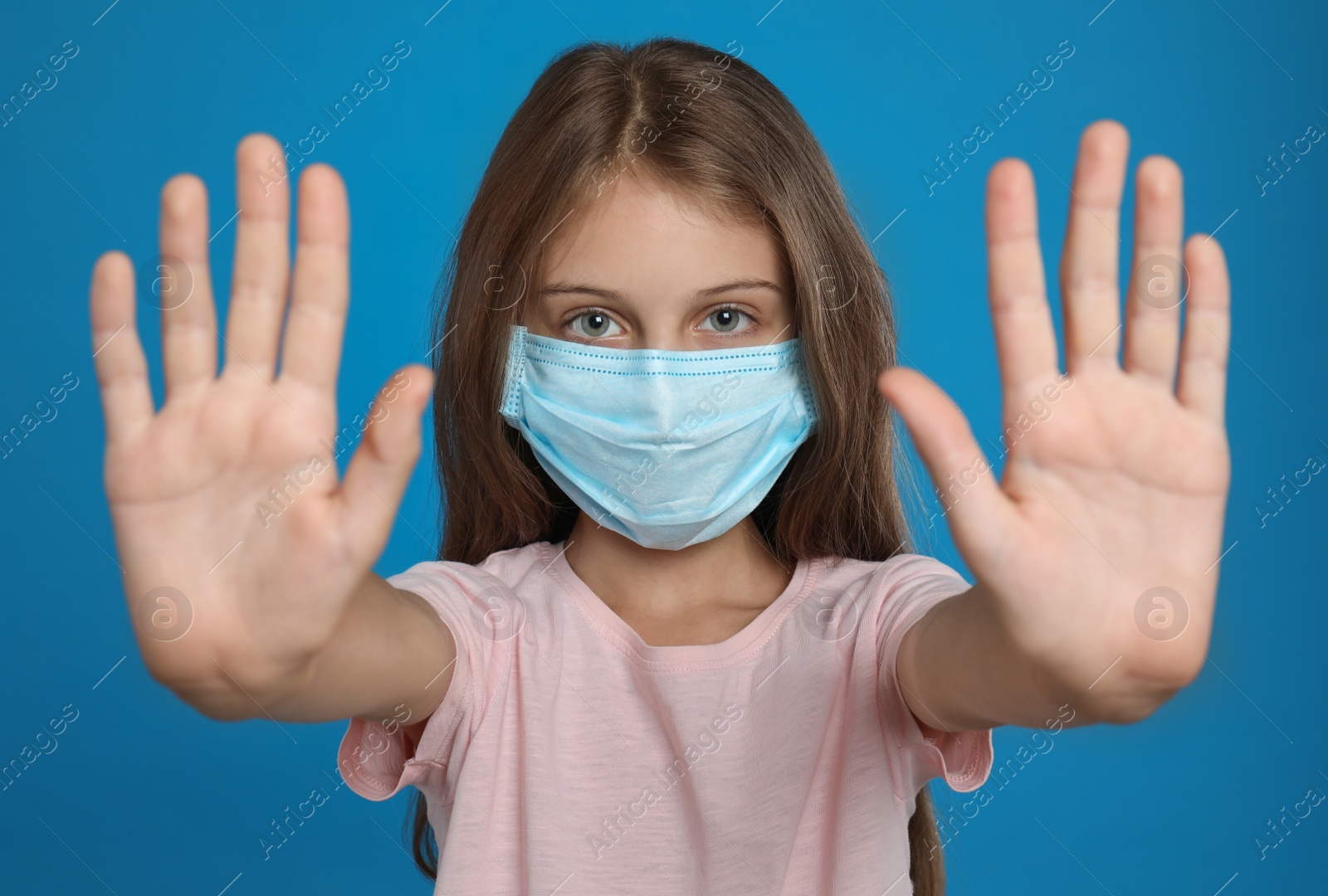 Photo of Little girl in protective mask showing stop gesture on blue background. Prevent spreading of coronavirus