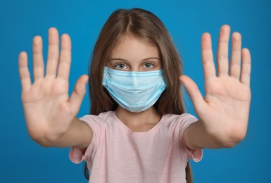 Photo of Little girl in protective mask showing stop gesture on blue background. Prevent spreading of coronavirus