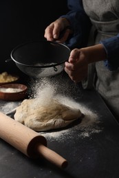 Woman sprinkling flour over dough at black table, closeup