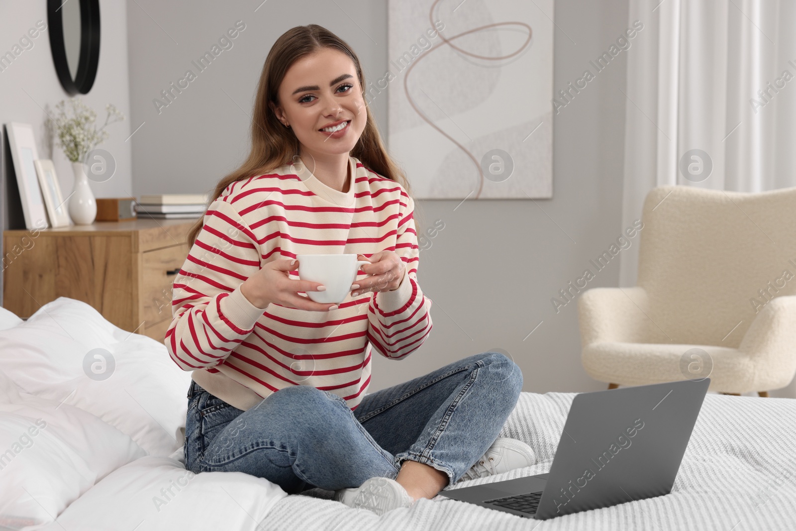 Photo of Happy woman with cup of drink and laptop on bed in bedroom