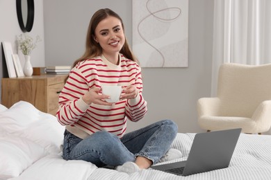 Happy woman with cup of drink and laptop on bed in bedroom