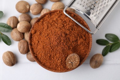 Photo of Nutmeg powder in bowl, seeds, grater and green branches on white table, flat lay