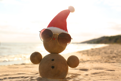 Photo of Snowman made of sand with Santa hat and sunglasses on beach near sea. Christmas vacation