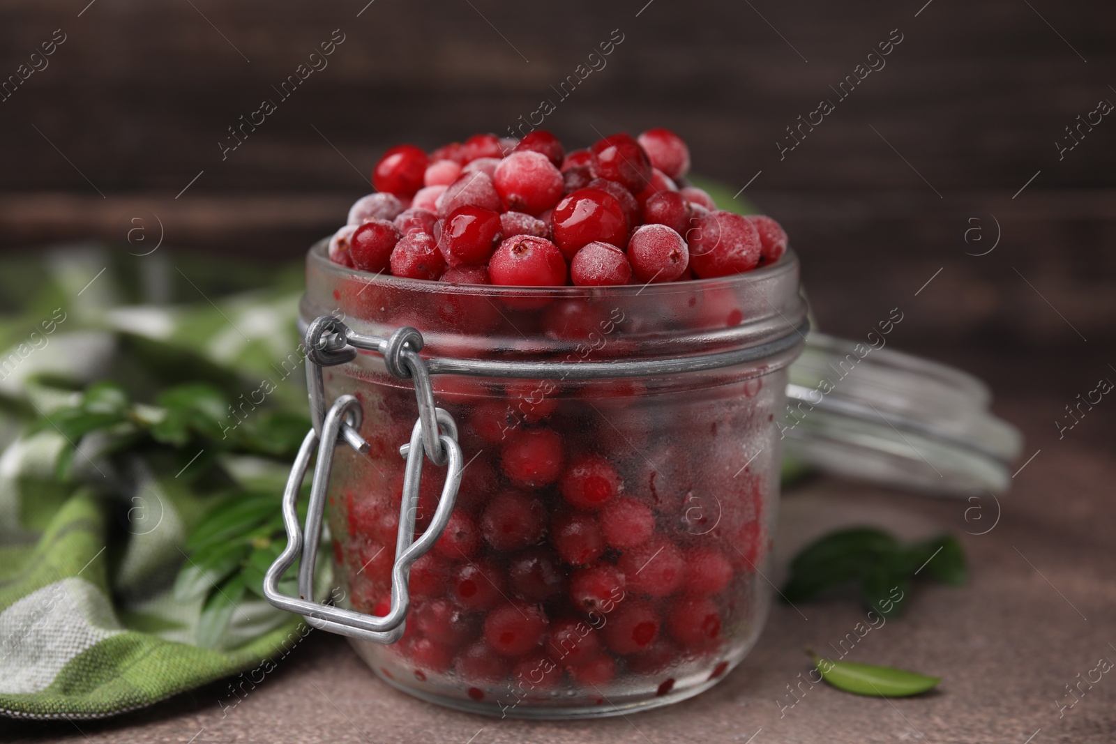 Photo of Frozen red cranberries in glass jar and green leaves on brown textured table, closeup