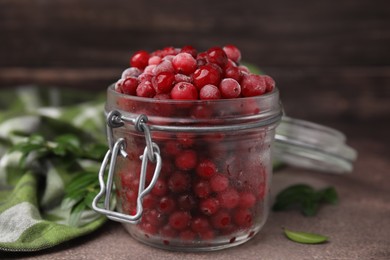 Photo of Frozen red cranberries in glass jar and green leaves on brown textured table, closeup
