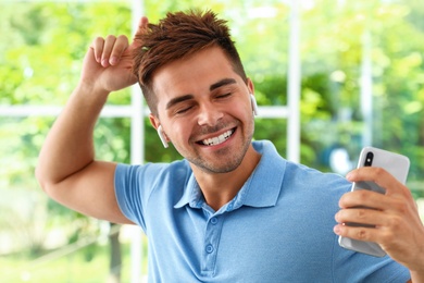 Happy young man with smartphone listening to music through wireless earphones indoors