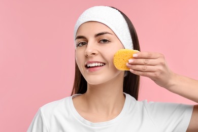 Photo of Young woman with headband washing her face using sponge on pink background