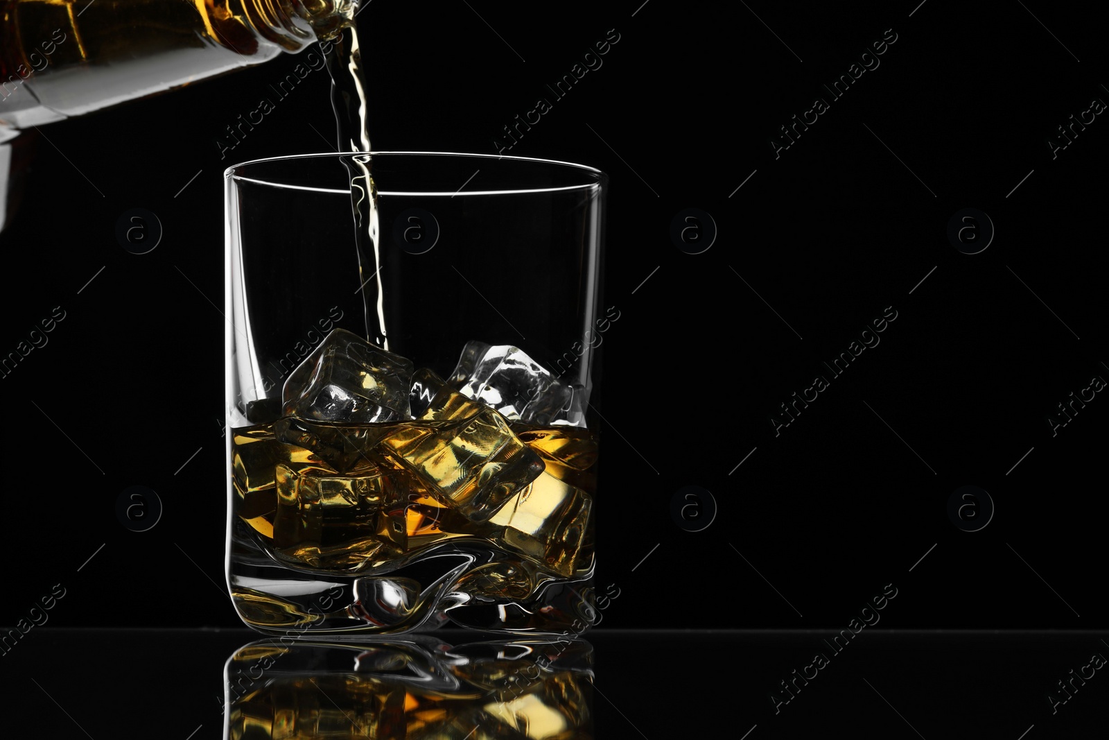 Photo of Pouring whiskey into glass with ice cubes at table against black background, space for text