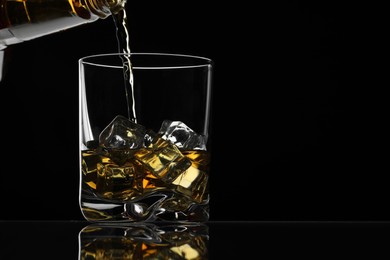 Photo of Pouring whiskey into glass with ice cubes at table against black background, space for text