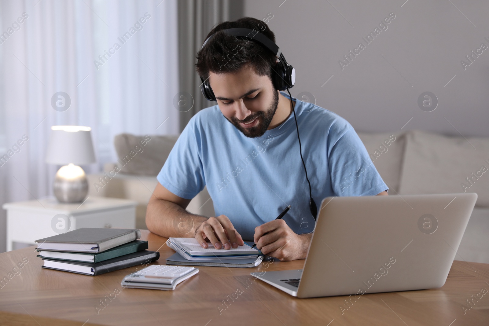 Photo of Young man writing down notes during webinar at table in room