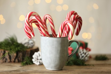 Photo of Christmas candy canes in cup on wooden table against blurred lights