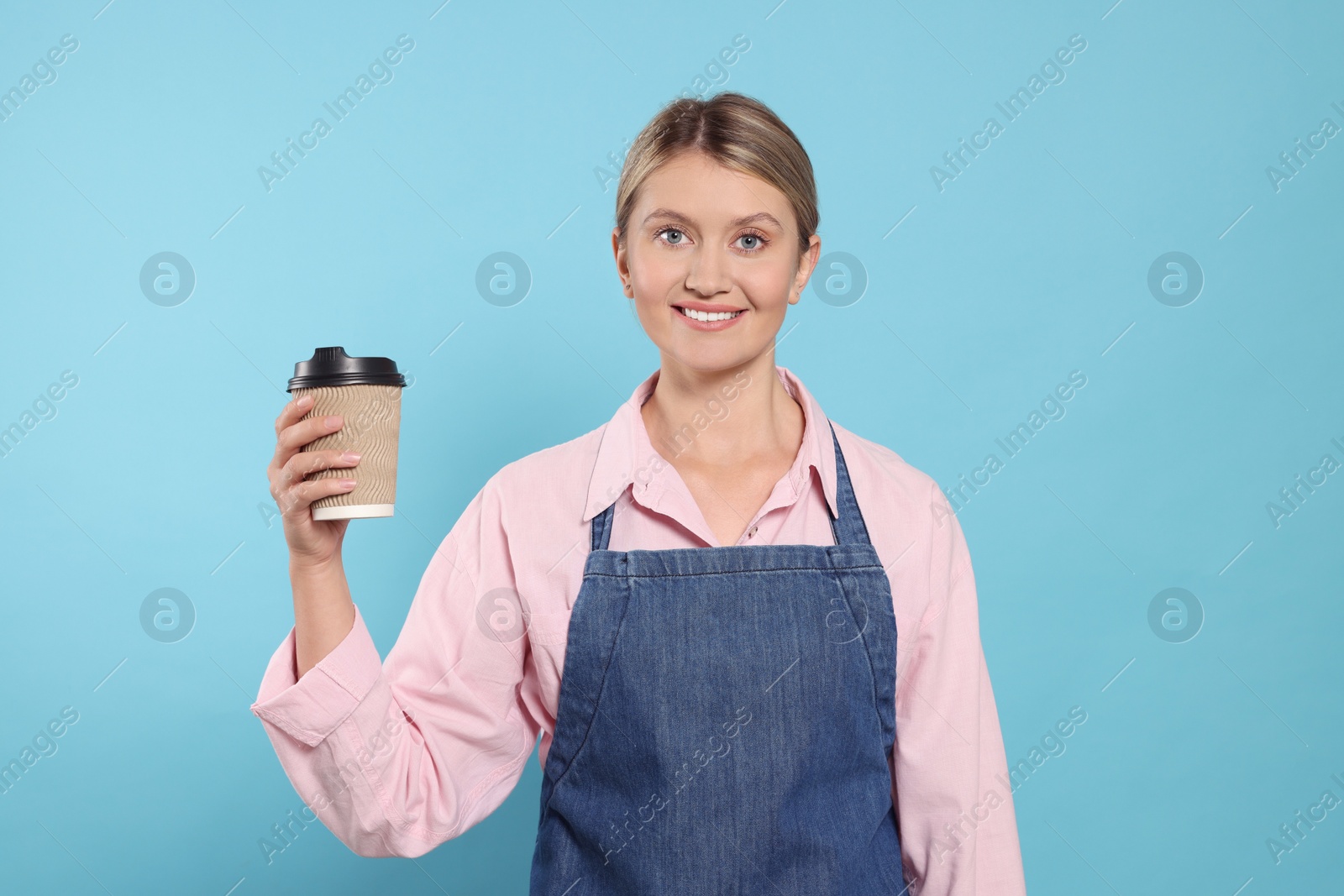 Photo of Beautiful young woman in denim apron with cup of coffee on light blue background