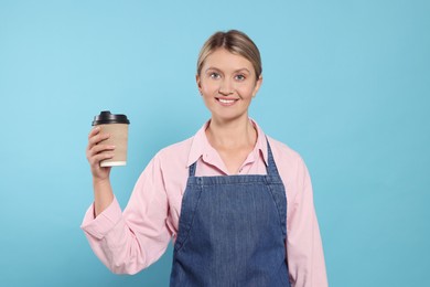 Beautiful young woman in denim apron with cup of coffee on light blue background