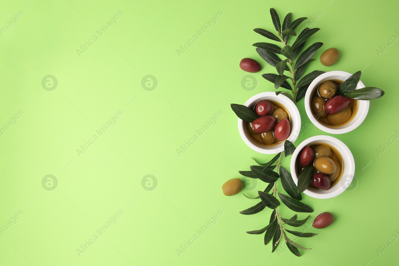 Photo of Bowls with different ripe olives and leaves on light green background, flat lay. Space for text