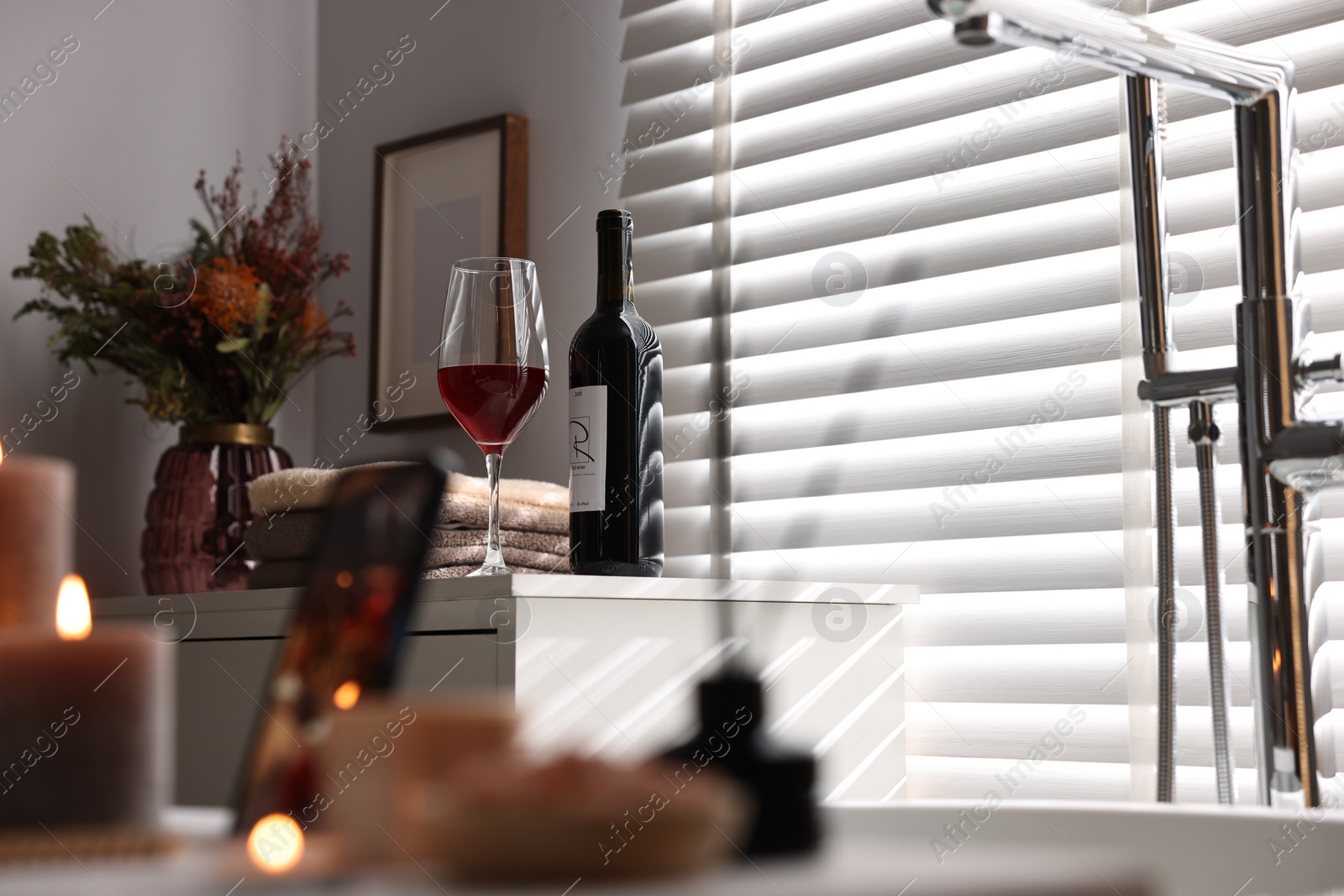 Photo of Bottle of wine, towels and flowers on chest of drawers in bathroom, selective focus