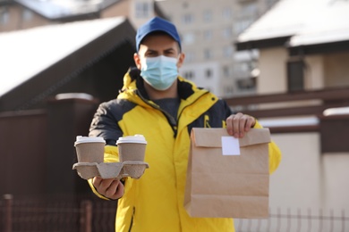 Photo of Courier in medical mask holding takeaway food and drinks near house outdoors, focus on paper cups. Delivery service during quarantine due to Covid-19 outbreak