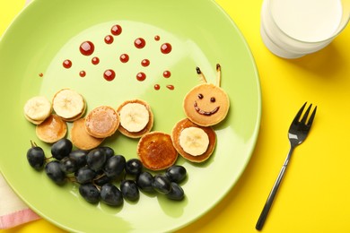 Photo of Creative serving for kids. Plate with cute caterpillar made of pancakes, grapes and banana on yellow background, flat lay