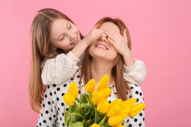 Photo of Daughter covering mother's eyes with her palms on pink background. Woman holding bouquet of yellow tulips