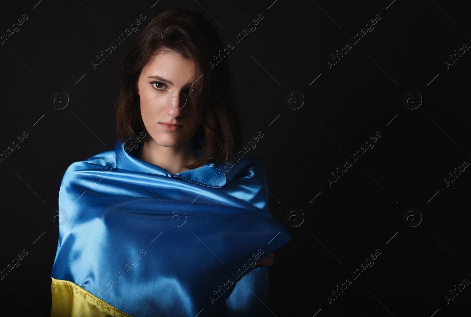 Photo of Sad woman wrapped in Ukrainian flag on black background. Space for text