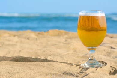 Glass of cold beer on sandy beach near sea. Space for text