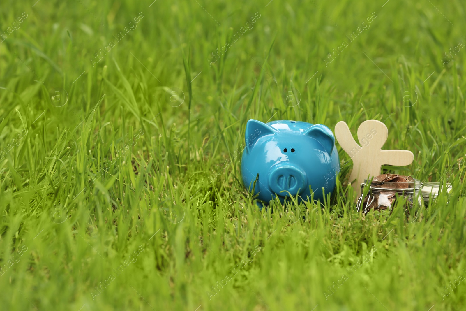 Photo of Piggy bank, wooden person and jar with coins on green grass in park. Space for text