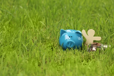 Piggy bank, wooden person and jar with coins on green grass in park. Space for text