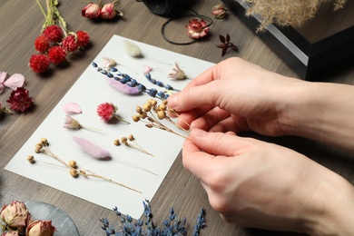 Photo of Woman making herbarium of dry flowers at wooden table, closeup