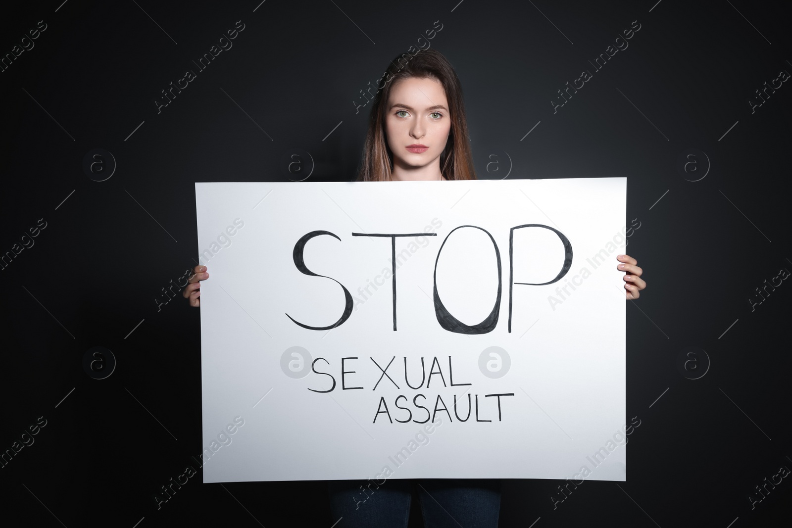 Photo of Young woman holding card with words STOP SEXUAL ASSAULT against dark background