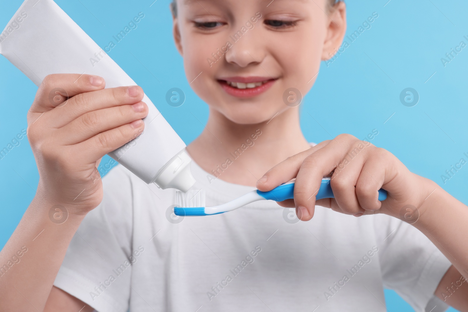 Photo of Happy girl squeezing toothpaste from tube onto toothbrush on light blue background, selective focus