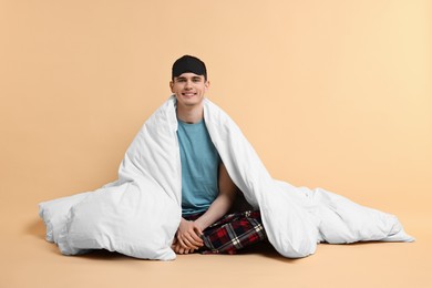 Happy man in pyjama and sleep mask wrapped in blanket on beige background