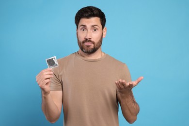 Confused man holding condom on light blue background. Safe sex