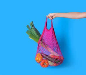 Photo of Woman holding net bag with vegetables on light blue background, closeup