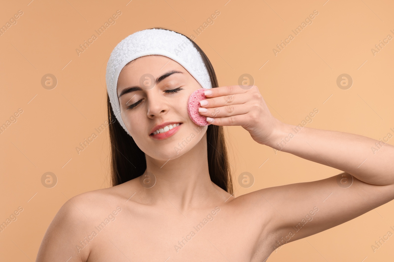 Photo of Young woman with headband washing her face using sponge on beige background