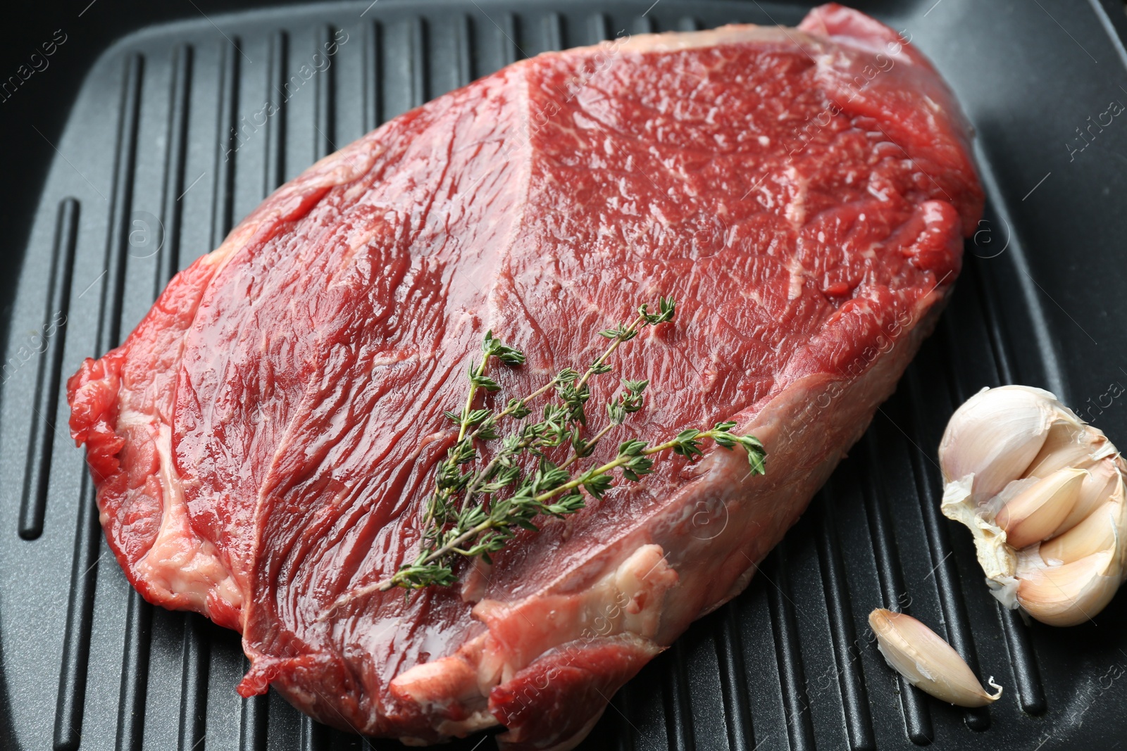 Photo of Grill pan with piece of raw beef meat, garlic and thyme, closeup