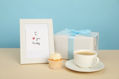 Happy Father's Day. Tasty cupcake, cup, card with phrase I Love You, Daddy in frame and gift box on beige table, closeup