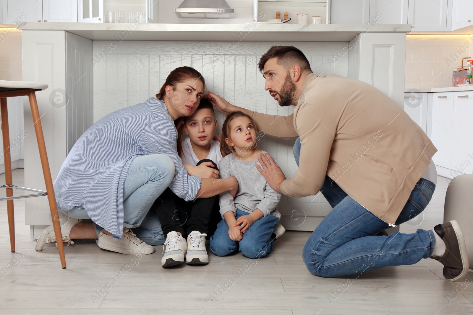 Photo of Scared parents with their children hiding under table in kitchen during earthquake