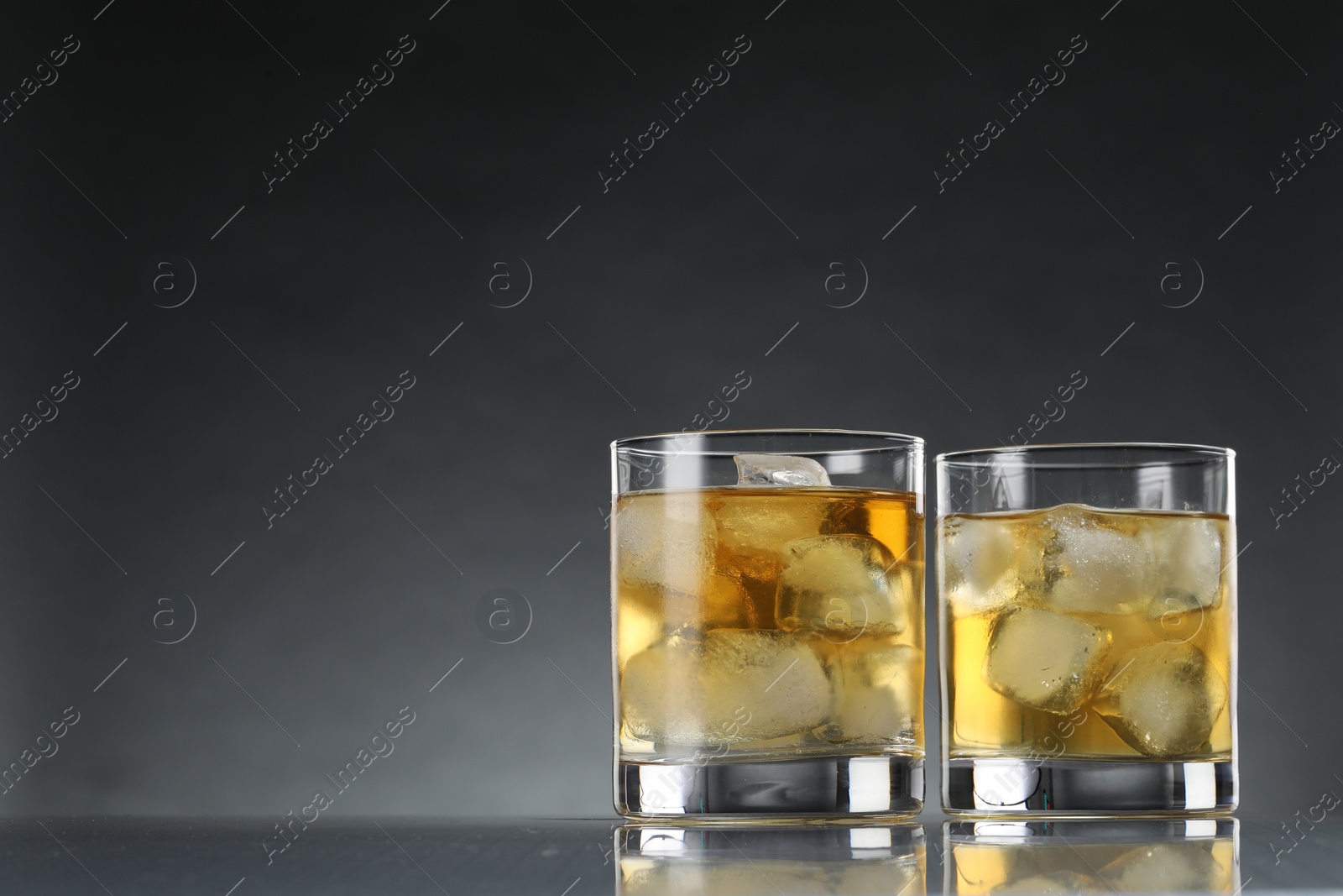 Photo of Tasty whiskey with ice in glasses on mirror table against dark background. Space for text