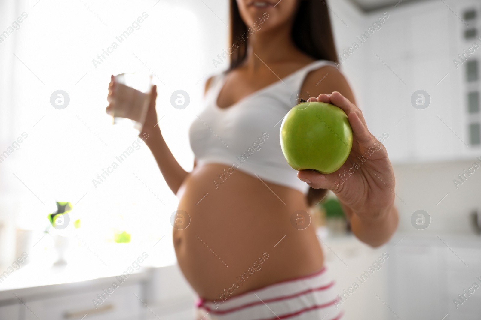 Photo of Young pregnant woman holding glass of water and apple in kitchen, focus on hand with fruit. Taking care of baby health